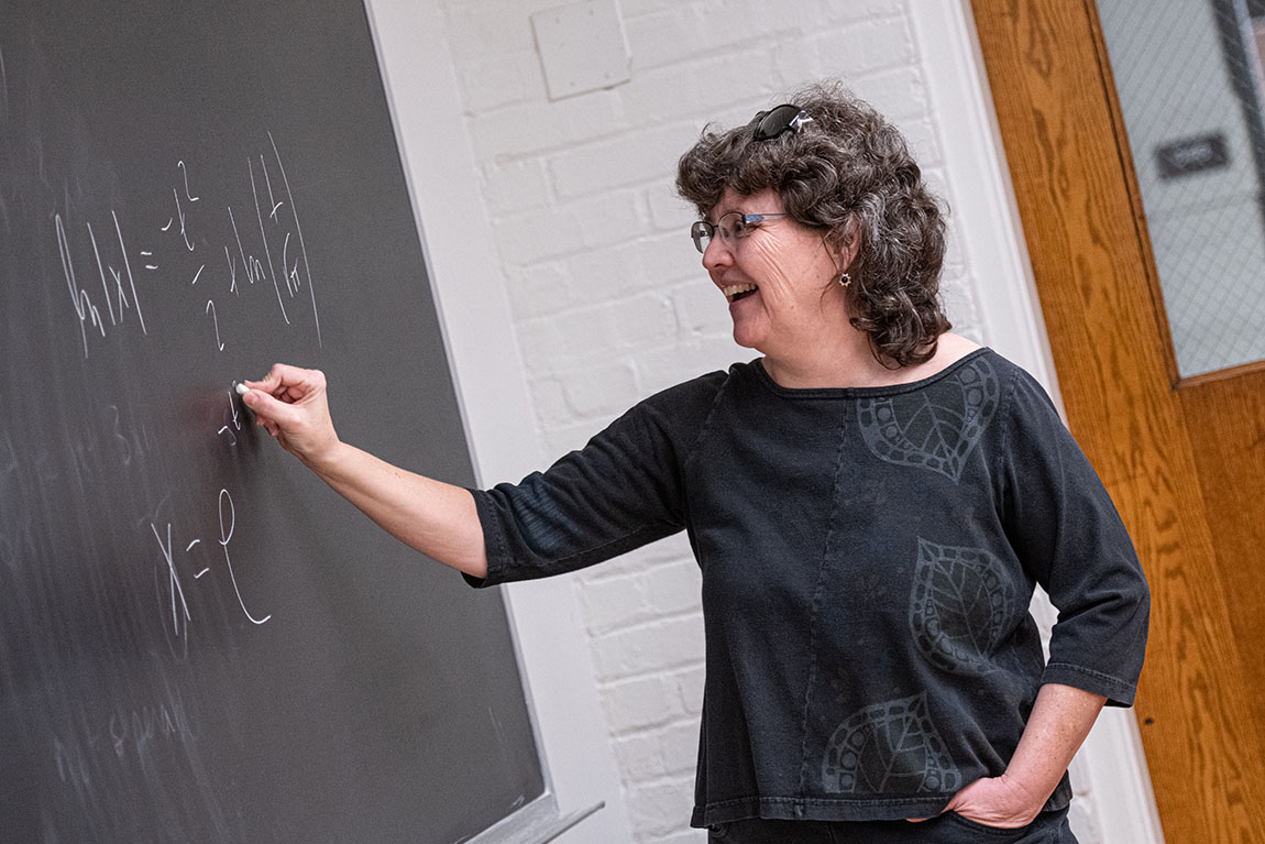 A college math professor with shoulder-length hair smiles as she writes equations on a chalkboard